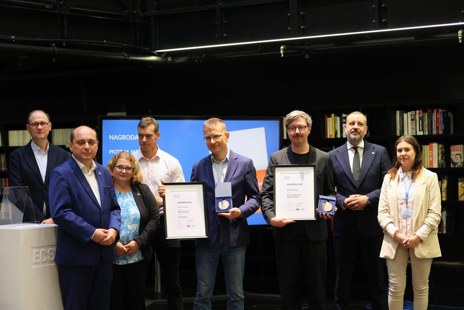 Kilka osób pozuje do zdjęcia podczas gali wręczenia nagród w konkursie wydawniczym. Dwóch mężczyzn trzyma dyplomy i medale. Gala odbywa się w nowoczesnych wnętrzach biblioteki.