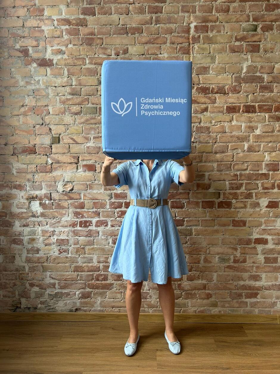 Zdjęcie stojącej kobiety, która na wysokości łowy trzyma prostokąt z napisem Gdański Miesiąc Zdrowia Psychicznego 