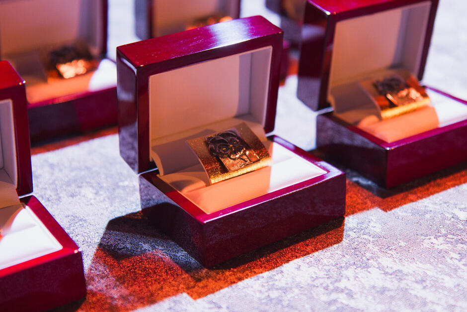 Na zdjęciu widać eleganckie, bordowe pudełka, w których znajdują się złote odznaczenia lub medale. Pudełka są otwarte i ustawione w rzędzie na stole pokrytym materiałem.