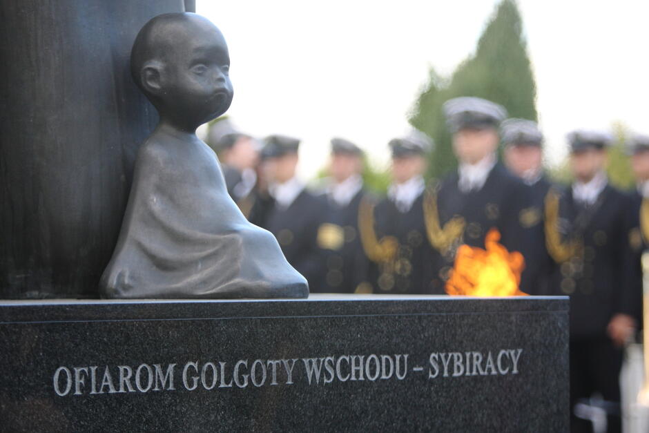 Cokół pomnika Golgota Wschodu. Widać siedzącą rzeźbę malutkiego dziecka - w tle płomienie znicza i oddział mężczyzn w mundurach Marynarki Wojennej RP 