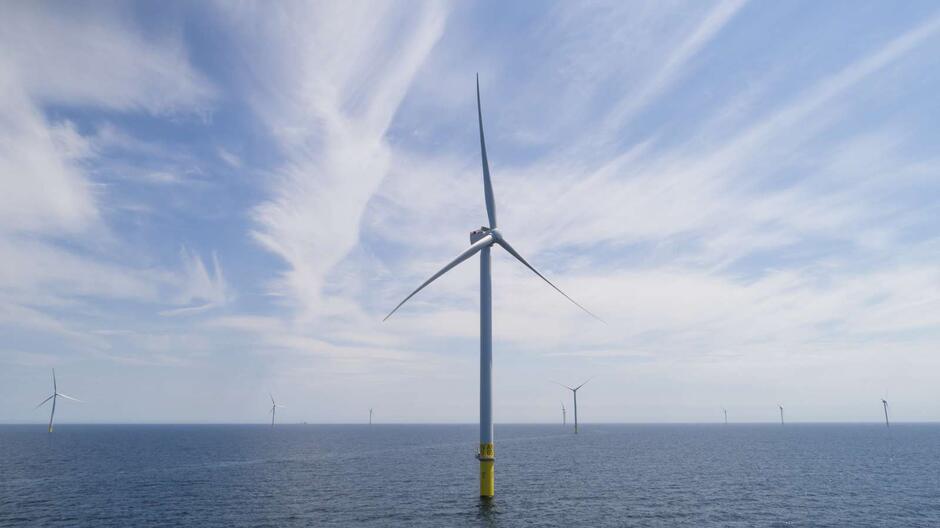 Na zdjęciu widać farmę wiatrową na morzu, gdzie wysokie turbiny wiatrowe stoją na solidnych fundamentach zanurzonych w wodzie. Na pierwszym planie dominuje pojedyncza turbina, a w tle rozciąga się szereg kolejnych, tworząc nowoczesny krajobraz energetyki odnawialnej.
