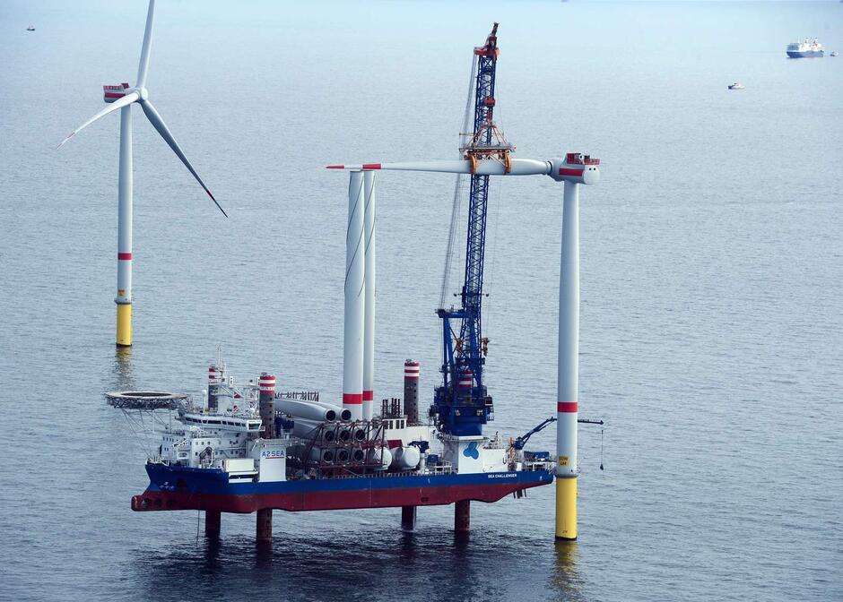 Na zdjęciu widać statek instalacyjny z dźwigiem montującym elementy turbiny wiatrowej na morzu. Obok znajduje się już ukończona turbina, a w tle widoczne są kolejne jednostki pływające.
