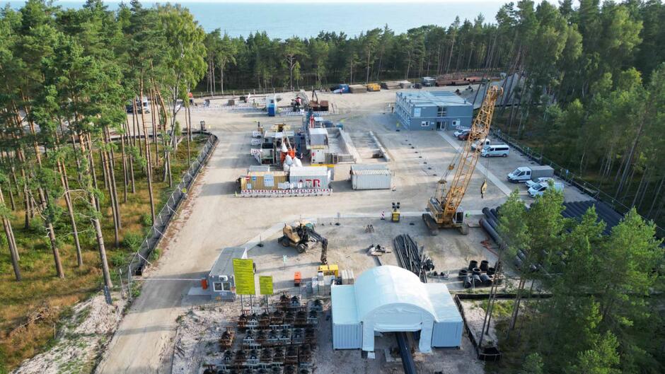 Zdjęcie przedstawia plac budowy zlokalizowany w pobliżu lasu i morza, gdzie prowadzone są prace związane z instalacją infrastruktury energetycznej. Na terenie widoczne są kontenery, dźwig, maszyny budowlane oraz elementy kabli i rur przygotowane do montażu.

