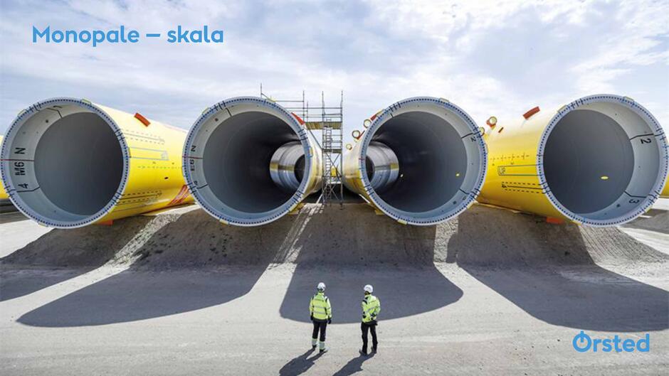 Na zdjęciu widać ogromne fundamenty typu monopale, które służą do osadzania morskich turbin wiatrowych. Ich imponującą skalę podkreślają stojący obok dwaj pracownicy w kaskach i odzieży ochronnej.
