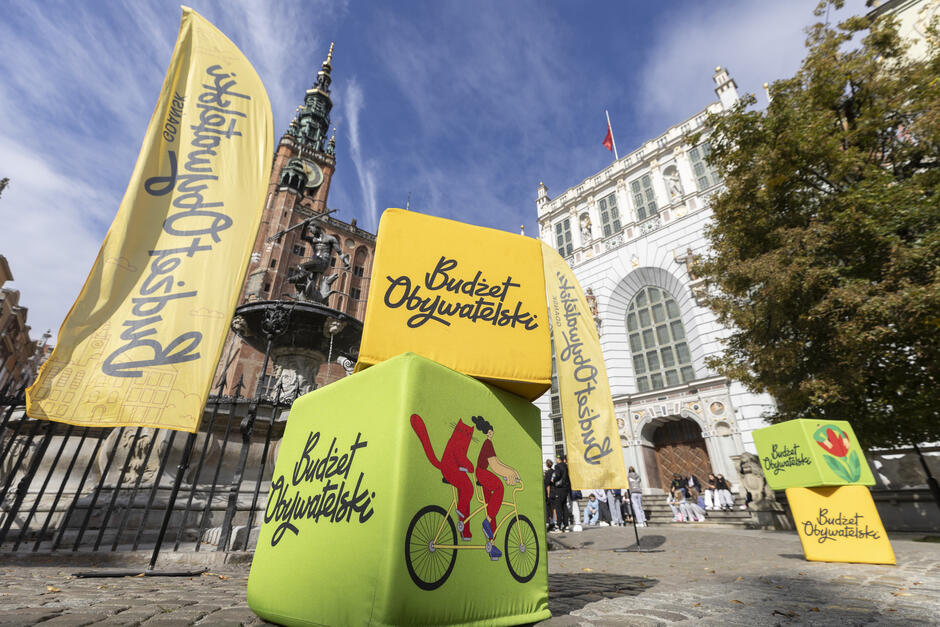 na zdjęciu dwie pluszowe kostki, jedna w kolorze zielonym druga żółtym, z kolorowymi grafikami, w tle obok żółte wysokie flagi z czarnym napisem budżet obywatelski