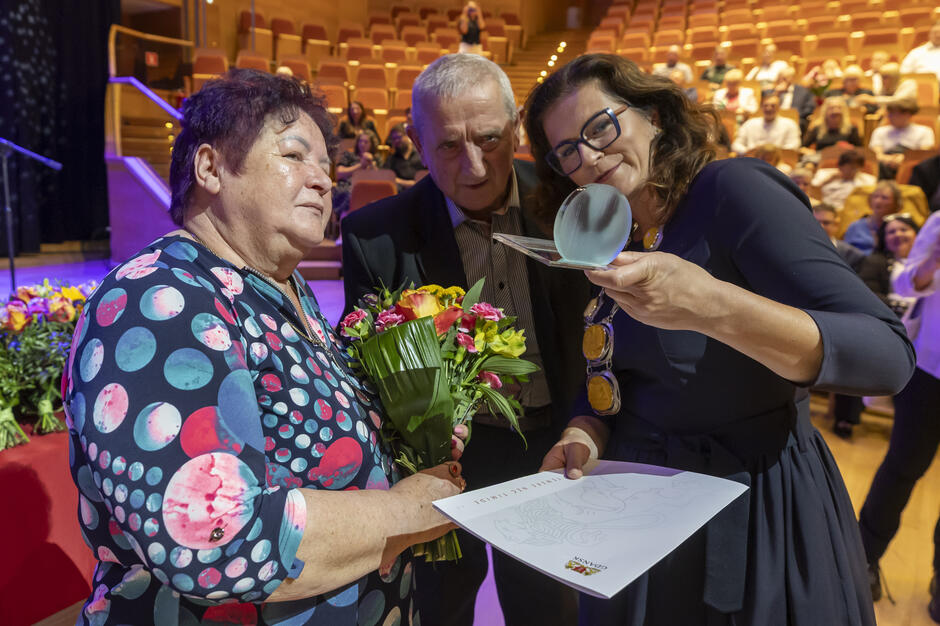 Kobieta wręcza parze starszych ludzi statuetkę w sali koncertowej.