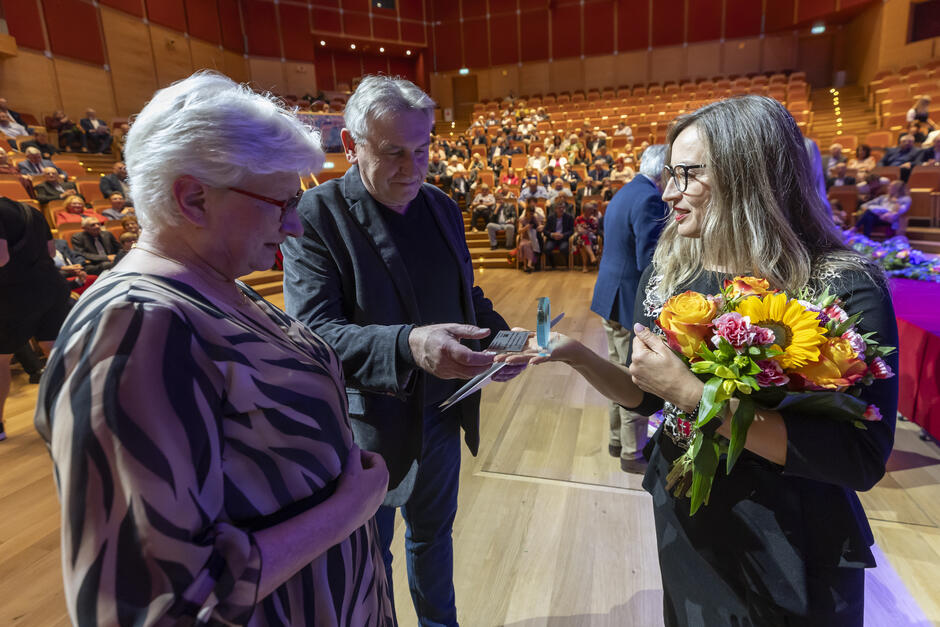 Kobieta wręcza statuetkę parze starszych osób w sali koncertowej.