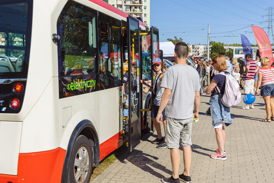 grupa ludzi stoi przed drzwiami małego autobusu. Na szybie pojazdu widoczny napis elektryczny . W tle widoczne kolorowe flagi i duże zgromadzenie ludzi