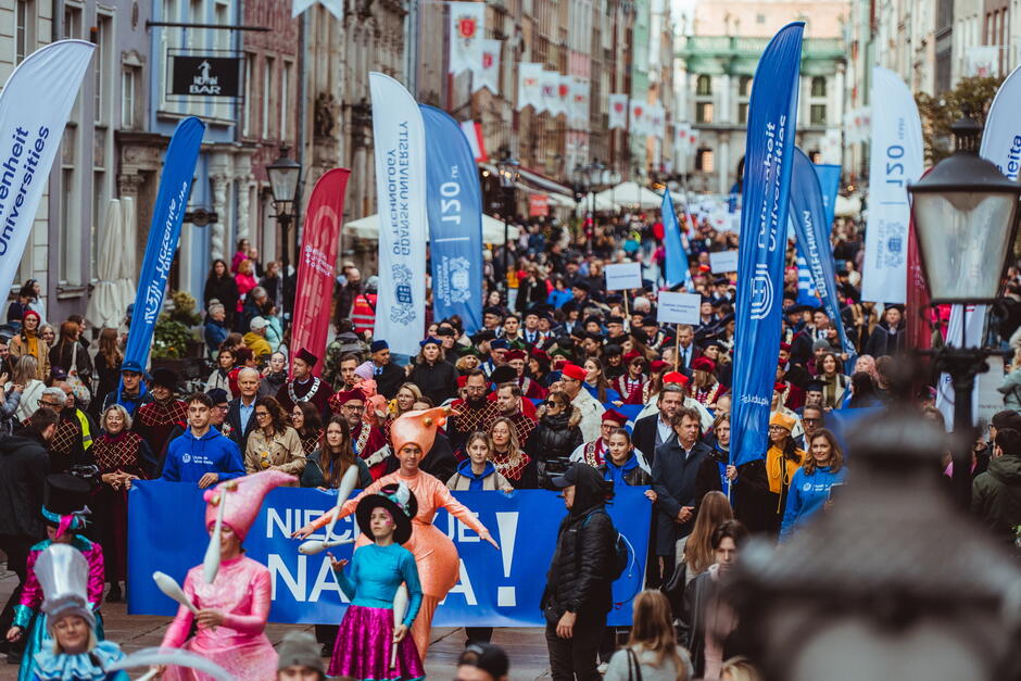 Ulicą starego miasta idzie parada społeczności akademickiej.