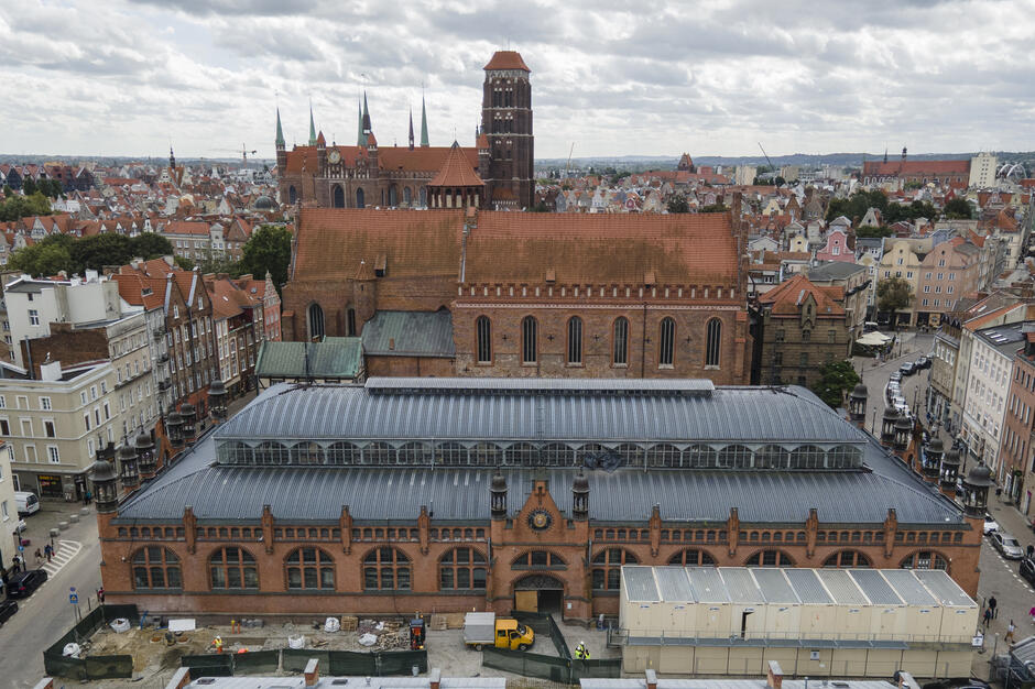 Na zdjęciu widać Halę Targową w Gdańsku na tle monumentalnej Bazyliki Mariackiej oraz zabytkowej zabudowy Głównego Miasta. Charakterystyczna ceglana architektura i stalowo-szklany dach hali kontrastują z gotyckimi murami kościoła oraz malowniczymi kamienicami wokół. 