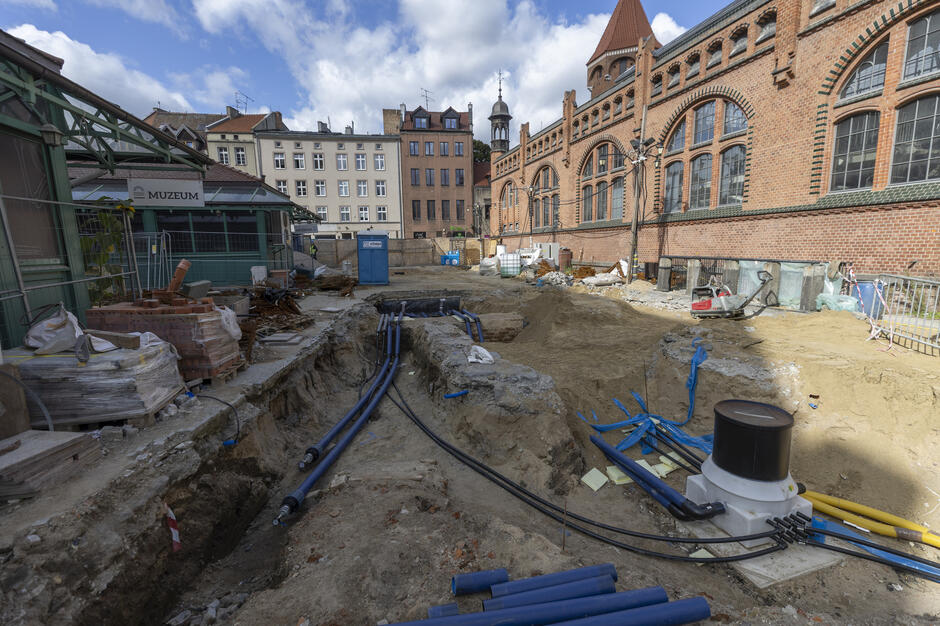 Na zdjęciu widać teren budowy przy Hali Targowej w Gdańsku, gdzie prowadzone są prace ziemne i instalacyjne, widoczne są wykopy z rurami i kablami. W tle znajduje się ceglana elewacja hali oraz otaczające ją kamienice. 