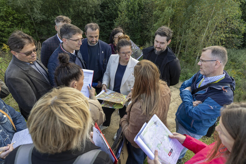 na zdjęciu grupa osób skupionych w kręgu, stoją blisko, dyskutują kobieta trzyma mapę papierową