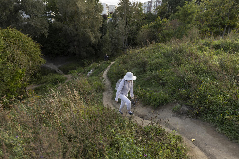 na zdjęciu kobieta w jasnym kapeluszu spaceruje po zielonym wzgórzu wydeptaną ścieżką