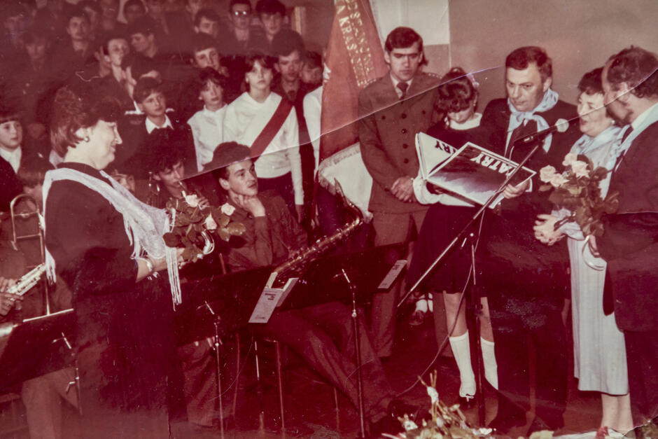 stara kolorowa fotografia przedstawia odświętnie ubranych uczniów ze sztandarem oraz nauczycieli w trakcie uroczystej akademii