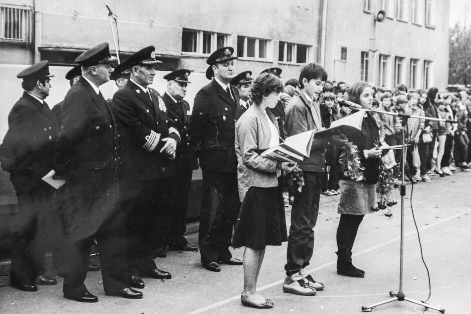 czarnobiałe zdjęcie przedstawia grupę młodzieży stojącą przy mikrofonie. W tle widoczny szpaler ludzi w marynarskich mundurach