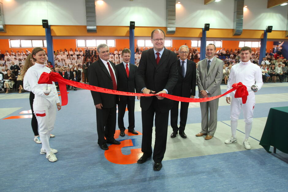 elegancko ubrany mężczyzna przecina nożyczkami wstęgę otoczony innymi równie elegancko ubranymi ludźmi. Za nimi widać wnętrze dużej sportowej hali 