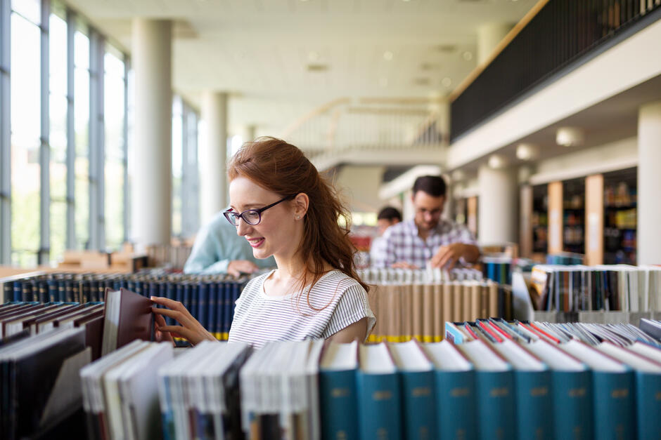 Na zdjęciu widać młodą kobietę w okularach, która przegląda książki na półce w bibliotece. W tle inni ludzie również wybierają książki, a wnętrze jest jasne i przestronne dzięki dużym oknom.