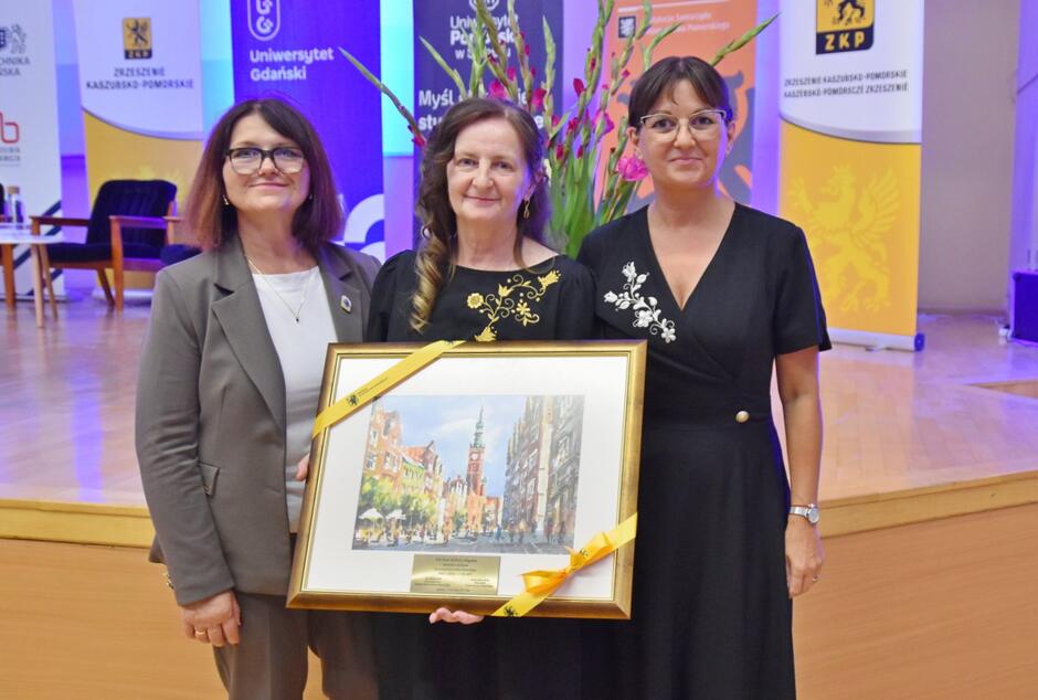 Na zdjęciu stoją trzy kobiety, z których środkowa trzyma oprawiony obraz przedstawiający fragment gdańskiej starówki. Wszystkie są uśmiechnięte i elegancko ubrane, a w tle widać bannery organizatorów wydarzenia oraz dekoracyjne kwiaty.