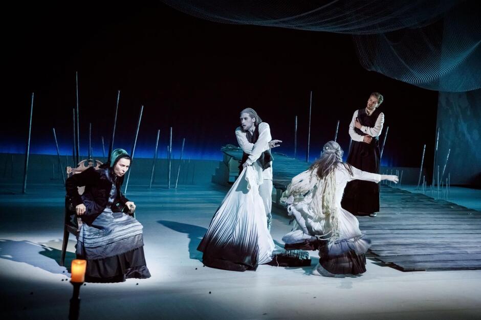 Na zdjęciu widać scenę teatralną z czterema aktorami ubranymi w kostiumy stylizowane na historyczne lub ludowe, w odcieniach czerni, bieli i szarości. Tło jest minimalistyczne, z drewnianym pomostem, wbitymi kijami i siatką zawieszoną nad sceną, co tworzy nastrojową, nieco mroczną atmosferę.