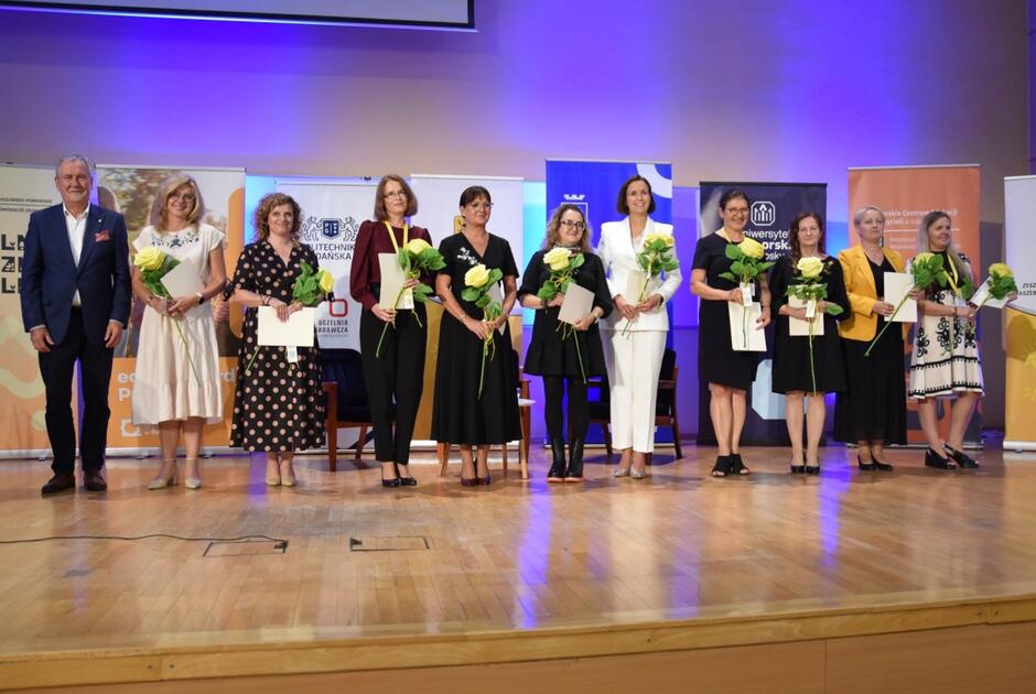 Na zdjęciu widać grupę osób stojących na scenie, z których większość to kobiety trzymające bukiety kwiatów i dyplomy. W tle widoczne są roll-upy różnych instytucji, a całość ma charakter oficjalnej uroczystości wręczenia wyróżnień.