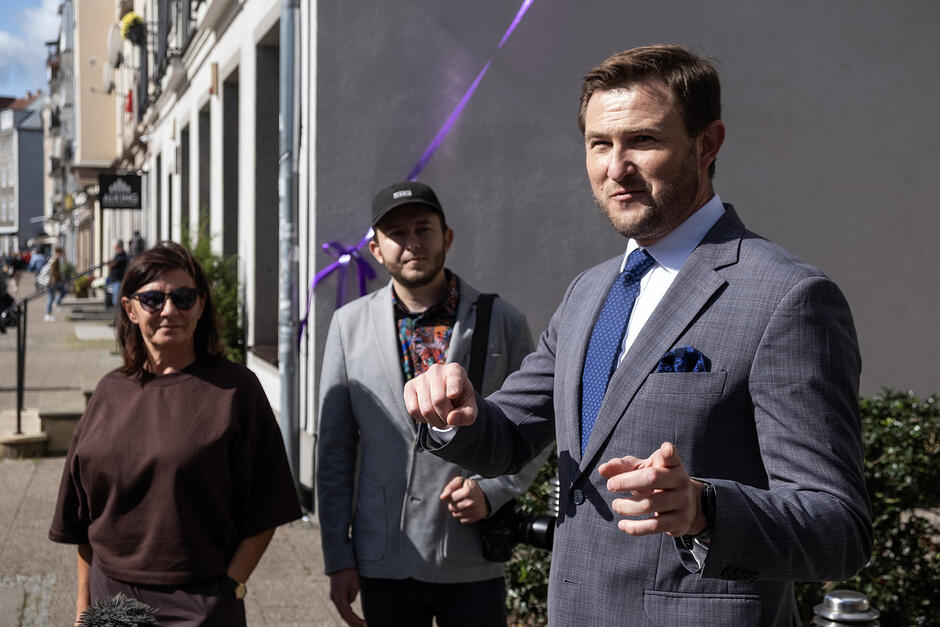 Dwóch mężczyzn i kobieta stoją na ulicy. Mężczyzna przemawia.