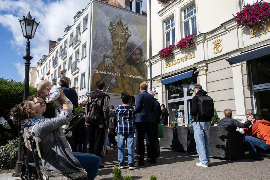 Tłum ludzi zebrany pod kamienica, na której widać mural z dwoma mężczyznami i figurą króla.