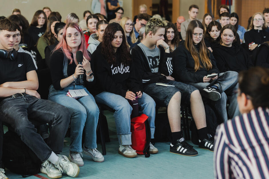 Na zdjęciu widać grupę młodzieży siedzącą w pierwszym rzędzie podczas spotkania, a jedna z uczennic z różowymi włosami trzyma mikrofon, przygotowując się do zadania pytania. Pozostali uczniowie obok niej uważnie słuchają lub trzymają w rękach notatki i książki.
