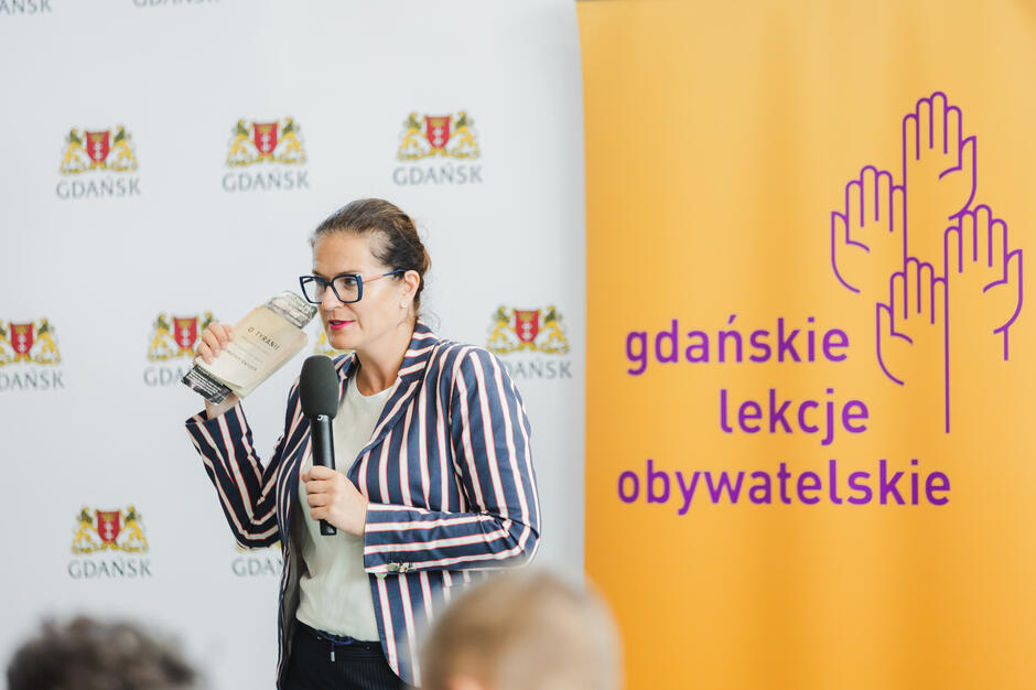 Na zdjęciu kobieta w marynarce w paski trzyma mikrofon oraz książkę „O tyranii” Timothy’ego Snydera, prowadząc wystąpienie. Obok niej znajduje się baner z napisem „gdańskie lekcje obywatelskie”, co wskazuje na edukacyjny charakter wydarzenia. 