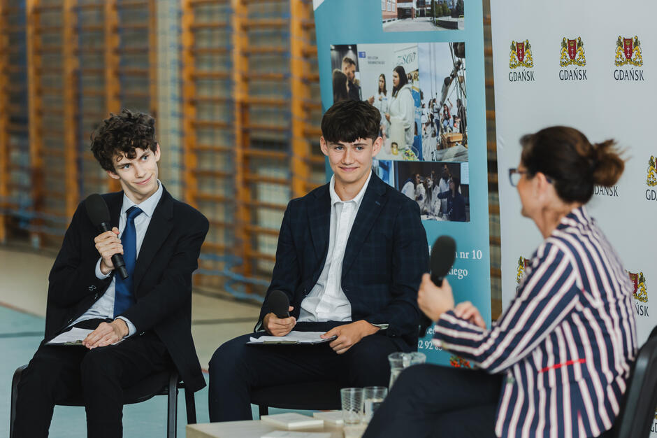 Na zdjęciu dwóch młodych chłopców w garniturach uczestniczy w rozmowie, trzymając mikrofony i notatki, co sugeruje udział w debacie lub panelu dyskusyjnym. Naprzeciwko nich siedzi kobieta w marynarce w paski, a w tle widoczne są banery z herbem Gdańska i informacjami o wydarzeniu. 