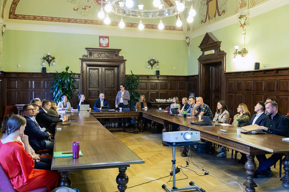 Kilkanaście osób siedzi przy stole ustawionym w podkowę w reprezentacyjnej sali.