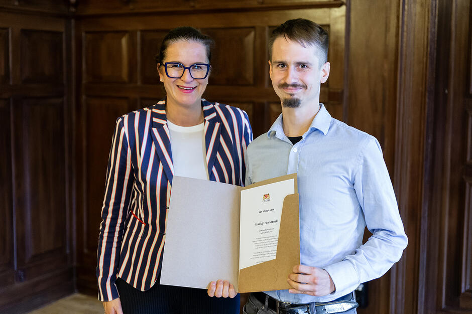 Kobieta z mężczyzną pozują do zdjęcia w reprezentacyjnej sali na tle drewnianej ściany. Mężczyzna pokazuje dokument.