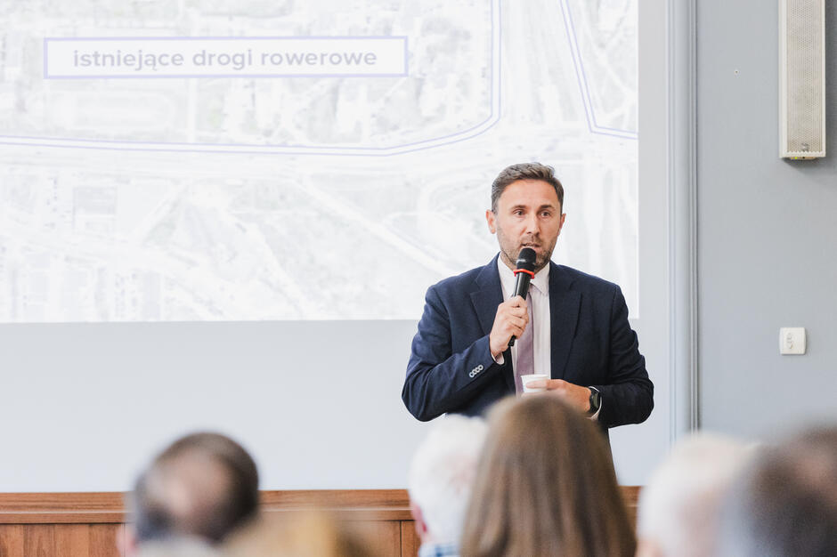 na zdjęciu mężczyzna w średnim wieku, w ciemnej marynarce, mówi do mikrofonu, przed nim widać od tyłu głowy siedzących osób