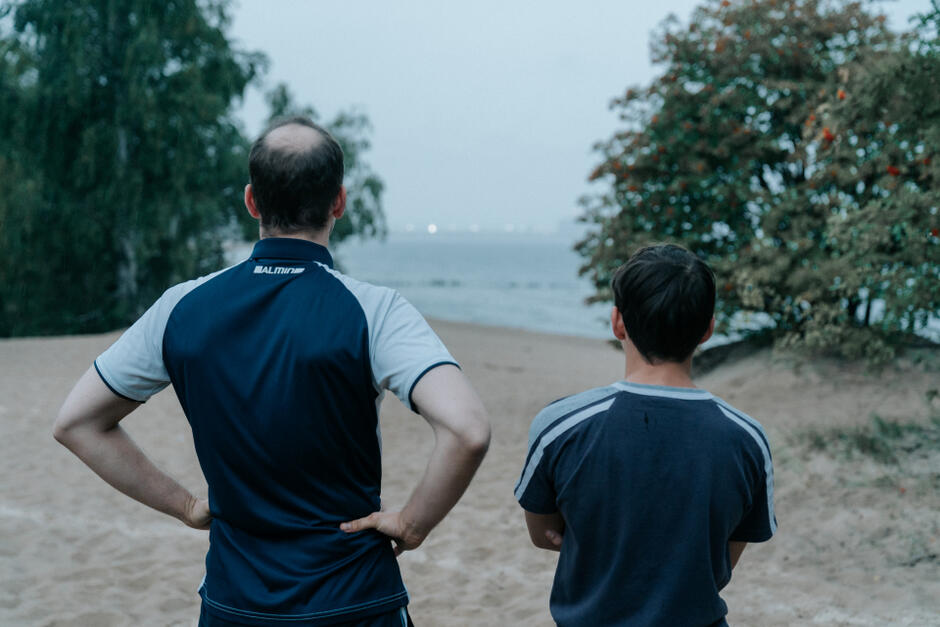 Dwóch chłopców stoi tyłem, patrząc w stronę morza z piaszczystej plaży. Sylwetki w sportowych koszulkach sugerują chwilę zadumy lub rozmowy.