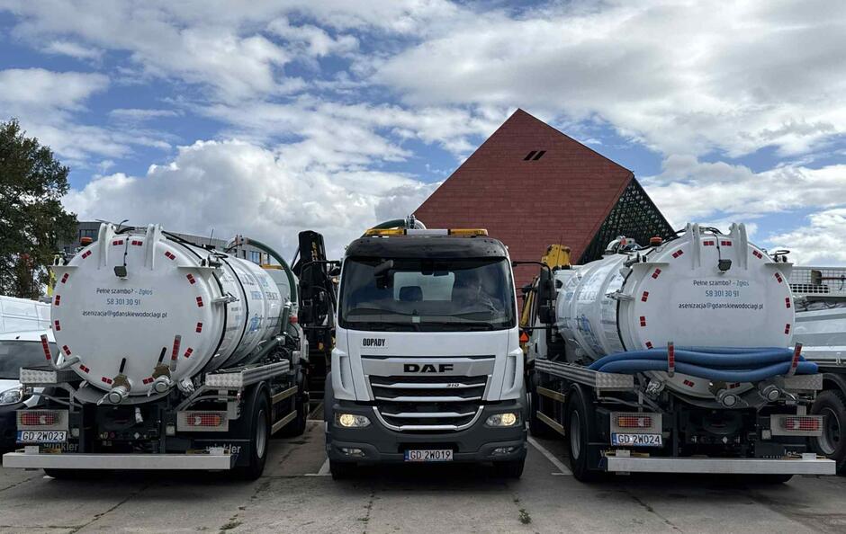 Trzy specjalistyczne pojazdy asenizacyjne stoją obok siebie na placu, a w tle widać charakterystyczny budynek Muzeum II Wojny Światowej w Gdańsku. Na beczkach umieszczono dane kontaktowe do zgłaszania wywozu nieczystości.