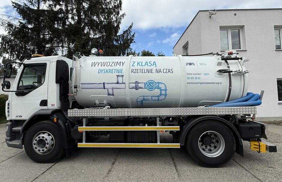 Biały samochód asenizacyjny z dużym zbiornikiem zaparkowany jest przy budynku i drzewach. Na boku widnieje hasło reklamowe „Wywozimy dyskretnie. Z klasą, rzetelnie, na czas” wraz z grafiką rury i zaworu.