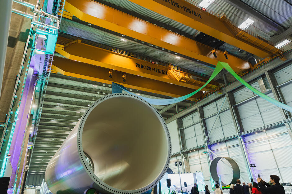 Na zdjęciu widać ogromną halę przemysłową, w której znajduje się wielki segment rury lub wieży turbiny wiatrowej. Nad całą przestrzenią zawieszony jest żółty suwnicowy dźwig, a w tle zgromadzili się ludzie oglądający prezentację.
