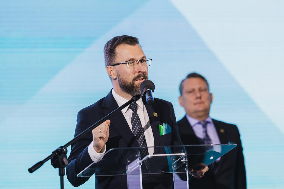 Na zdjęciu mężczyzna w garniturze przemawia przy mównicy z mikrofonem, gestykulując dłonią, co podkreśla wagę jego słów. W tle stoi inny mężczyzna w garniturze, słuchający wystąpienia.
