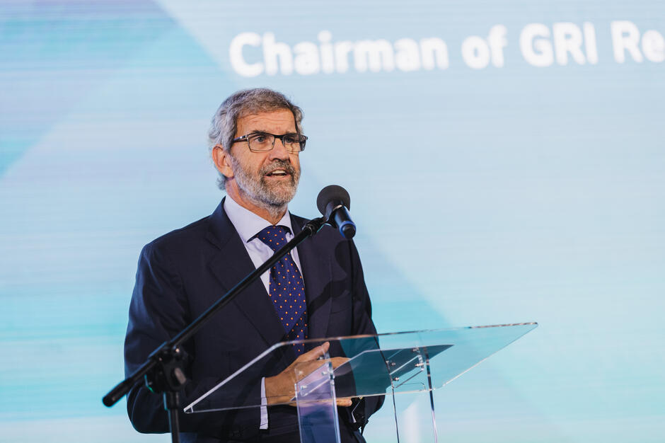 Na zdjęciu starszy mężczyzna w garniturze przemawia przy mównicy z mikrofonem, trzymając notatki w dłoni. W tle na ekranie widać fragment napisu „Chairman of GRI…”, co sugeruje jego rolę przewodniczącego organizacji.

