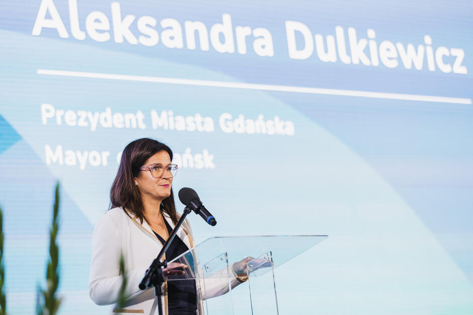 Na zdjęciu Aleksandra Dulkiewicz, prezydent Gdańska, przemawia przy mównicy z mikrofonem. W tle na dużym ekranie wyświetlone jest jej imię, nazwisko oraz funkcja w języku polskim i angielskim.
