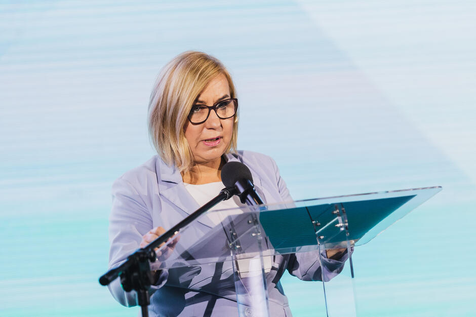 Na zdjęciu kobieta w jasnym garniturze i okularach przemawia przy mównicy z mikrofonem, trzymając notatki. Za nią widać jasne tło ekranu, które nadaje wystąpieniu oficjalny i uroczysty charakter.
