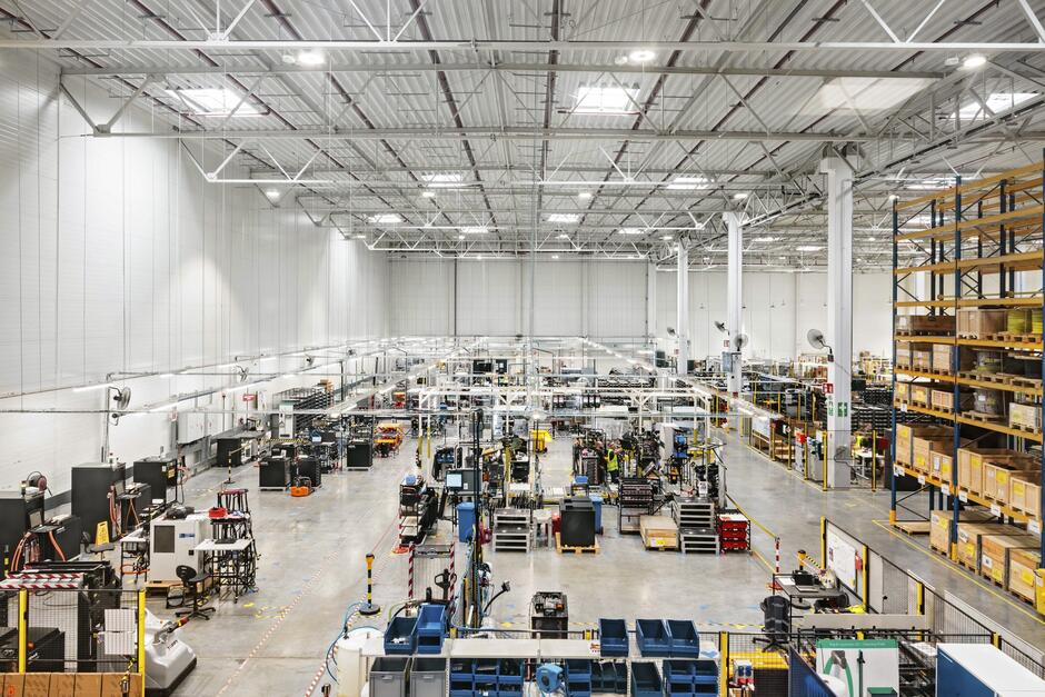 Na zdjęciu widoczna jest nowoczesna hala produkcyjna w Gdańsku, wypełniona maszynami, stanowiskami roboczymi oraz regałami z magazynowanymi materiałami. Przestrzeń jest jasna, uporządkowana i zaprojektowana tak, aby zapewnić sprawny przebieg procesów przemysłowych.
