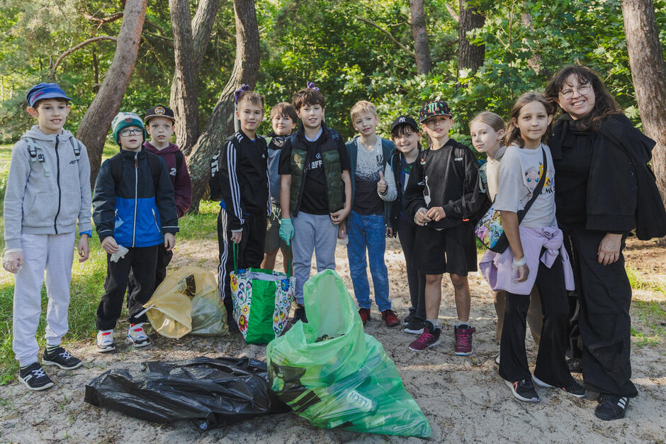Na zdjęciu grupa dzieci wraz z opiekunką pozuje w lesie podczas akcji sprzątania świata, trzymając wypełnione worki na śmieci. Wszyscy uśmiechają się do kamery, a kolorowe torby i worki pełne odpadów świadczą o ich zaangażowaniu w dbanie o środowisko.
