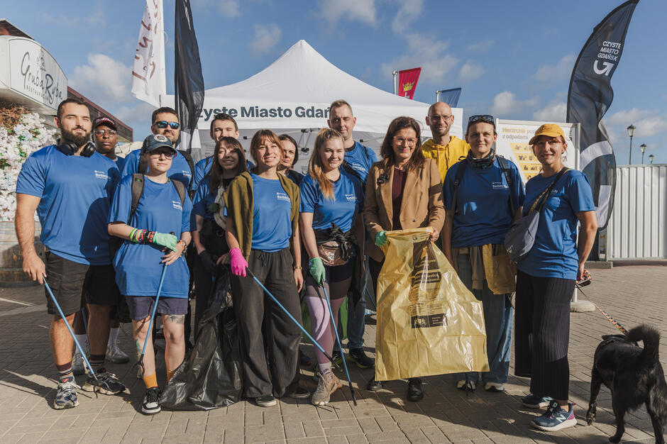 Na zdjęciu grupa wolontariuszy w niebieskich koszulkach pozuje wspólnie podczas akcji „Czyste Miasto Gdańsk”, trzymając worki na śmieci i chwytaki do odpadów. W tle widoczny jest biały namiot organizacyjny oraz flagi, co podkreśla oficjalny charakter wydarzenia. 