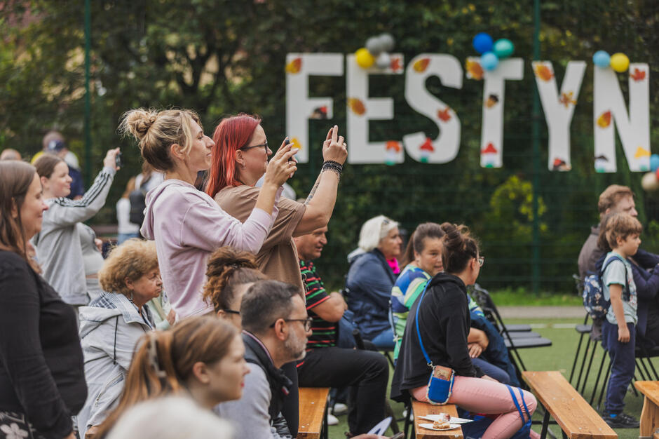 na zdjęciu dwie kobiety stoją nagrywają coś telefonami, w tle duży biały napis festyn