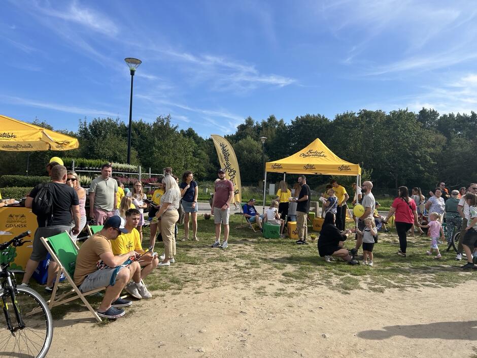 na zdjęciu piknik z żółtym namiotem, są też siedziska, część osób siedzi, inni stoją