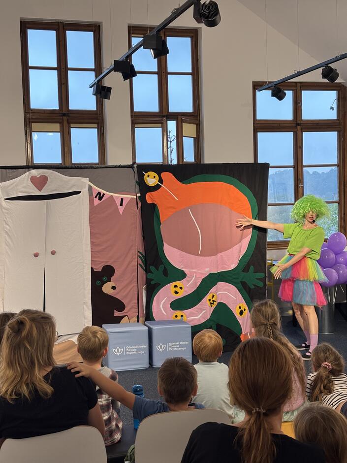 Na zdjęciu widać występ teatralny dla dzieci w jasnej sali z dużymi oknami. Przed grupą dzieci siedzących na krzesłach i podłodze stoi osoba w zielonej peruce, kolorowej tiulowej spódnicy i zielonej koszulce, wskazując na duży barwny baner przedstawiający schemat ludzkiego organizmu w formie bajkowej ilustracji. Obok znajduje się drugi baner z obrazkiem misia i sercem. Na podłodze stoją dwa siedziska z napisem „Gdański Miesiąc Psychoedukacyjny”. Dzieci skupione oglądają przedstawienie.