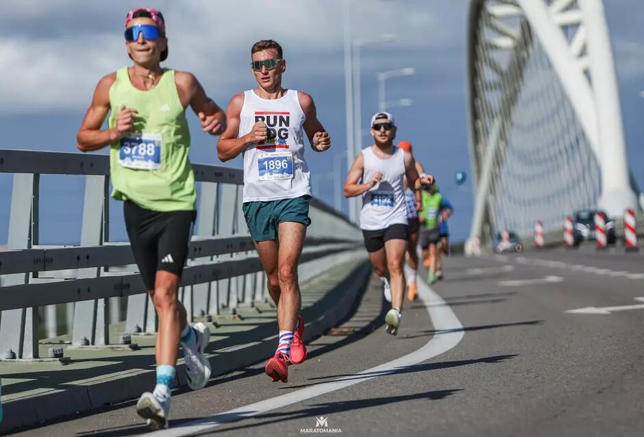 Kilku biegaczy w sportowych strojach pokonuje odcinek na moście w słoneczny dzień, każdy z numerem startowym przypiętym do koszulki. Na czele widać zawodnika w białej koszulce z napisem „RUN DOG”.