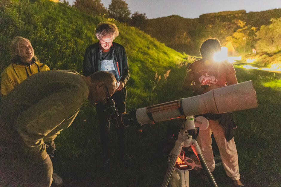 Na zdjęciu grupa osób obserwuje nocne niebo przez teleskop ustawiony na statywie, a wokół panuje atmosfera ciekawości i skupienia. Ciepłe światło w tle oraz zielone zbocze nadają scenie klimatyczny, przyjazny charakter. 