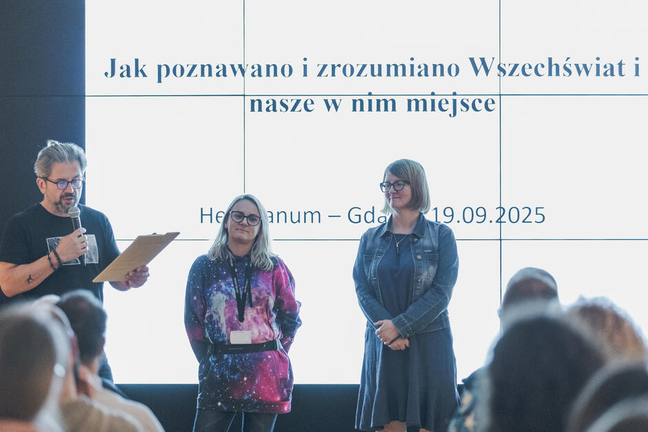  Trzy osoby stojące na scenie przed dużym ekranem z tekstem. Mężczyzna po lewej stronie trzyma mikrofon i wydaje się prezentować. Ma na sobie czarną koszulkę i okulary i stoi obok kobiety w różowo-fioletowej sukience z krawatem. Po prawej stronie stoi inna kobieta ze spłaszczonymi rękami i mężczyzna stoi za nią. Wszyscy patrzą w stronę publiczności, która siedzi w rzędach krzeseł skierowanych na scenę. Na ekranie za nimi znajduje się tekst Jak poznawano i zrozumiano Wszechświat i nasze w nim miejsce  