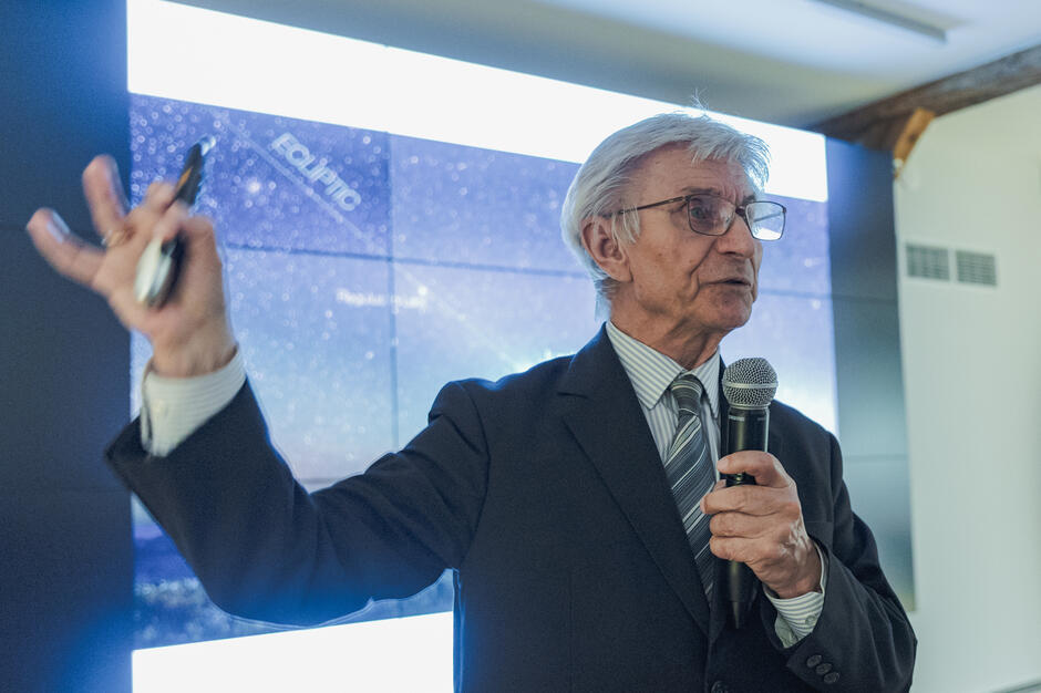 Na zdjęciu starszy mężczyzna w garniturze prowadzi wykład, trzymając w dłoni mikrofon i wskaźnik. W tle widoczny jest ekran z mapą nieba, co sugeruje, że prelekcja dotyczy astronomii lub kosmosu. 